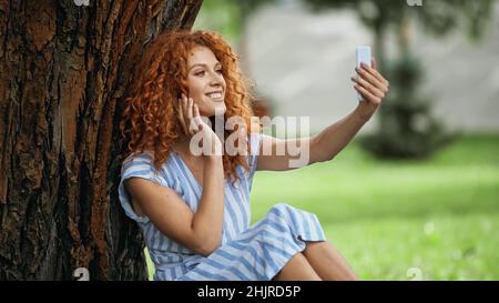 pleased redhead woman taking selfie while sitting under tree trunk Stock Photo