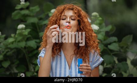 curly redhead woman blowing soap bubble in park Stock Photo