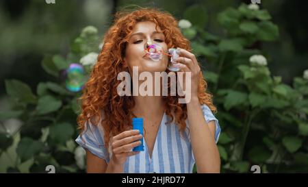 cheerful woman with red hair blowing soap bubbles in park Stock Photo