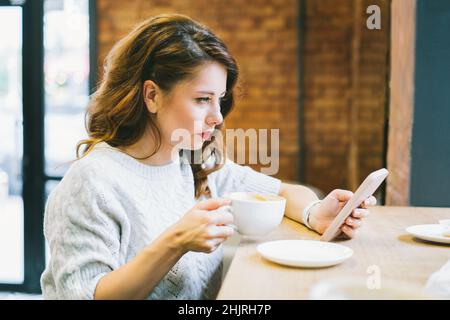 Concentrated young brunette female with cup of coffee in hands messaging on phone while sitting at table in comfortable cafe during lunch time break. Stock Photo