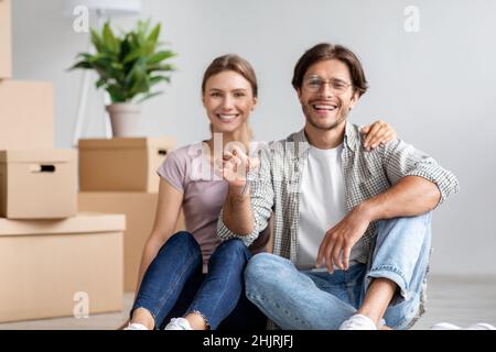 Satisfied caucasian young man in glasses and lady sit on floor show keys among cardboard box with stuff in room Stock Photo