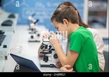 Boy looking through microscope near sitting teacher Stock Photo