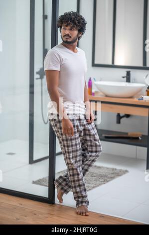 Calm thoughtful curly-haired barefoot male standing in the bathroom doorway Stock Photo