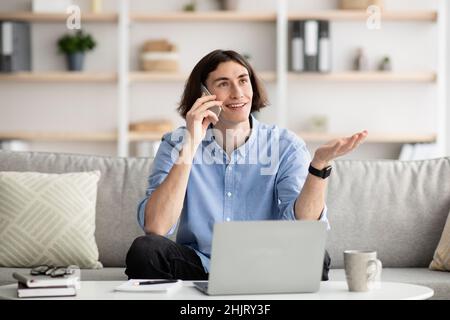 Positive young man working on laptop and having phone conversation, communicating with clients or business partners Stock Photo