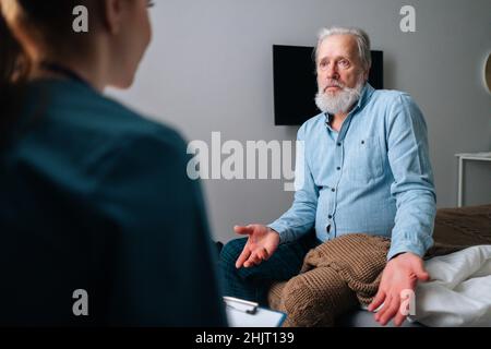 Close-up view from back of female doctor talking to frustrated senior male patient sitting on bed at nursing home. Stock Photo