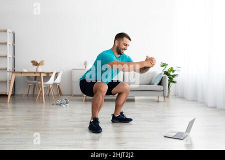 Happy millennial european guy doing leg exercises, squats and watching online lesson on pc Stock Photo
