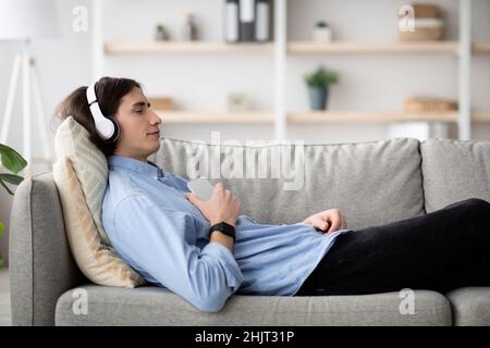 Rest and relax concept. Calm guy lying on sofa, listening to music, enjoying meditation for sleep in wireless headphones Stock Photo