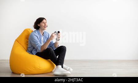 Young guy with joystick playing online video game, sitting in beanbag chair over light wall, panorama with free space Stock Photo