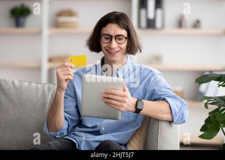 Online shopping concept. Happy young man holding debit credit card and using digital tablet, ordering food delivery Stock Photo