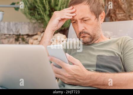 Worried freelancer and digital nomad working on smartphone and laptop computer during summer vacation holiday on apartment terrace in morning Stock Photo