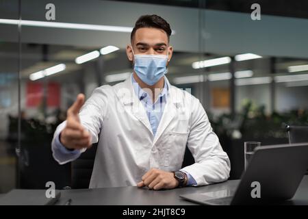 Doctor in face mask sitting at workdesk, greeting patient Stock Photo