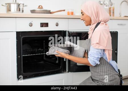 Beautiful Muslim Housewife Baking In Kitchen, Putting Mold With Pie To Oven Stock Photo