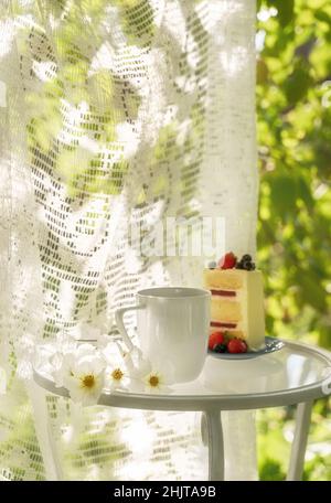 Beautiful porcelain coffee cup and slice of cake on glass table decorated with white flowers in summer garden in sunlight. Copy space. Summer drinks concept. Stock Photo
