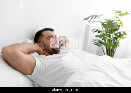 Happy caucasian young male in white t-shirt rests and relaxes, looks at empty space on bed in bedroom interior Stock Photo