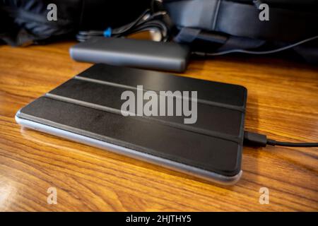 Angled view of an e-reader device in a black case, charging up on a wooden table, with backpacks in the background Stock Photo