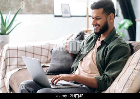 Positive handsome young indian man in casual clothes, sitting on the couch at home in the living room, using a laptop, browsing the Internet, messaging on social networks, looking at screen, smiles Stock Photo