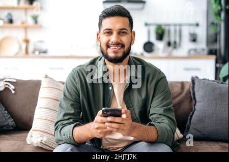 Happy handsome indian or arabian guy in casual wear, sits at home on sofa, using his smartphone, texting online, browsing social networks, messaging with friends, looks at camera, smiles friendly Stock Photo