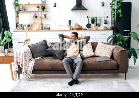 Satisfied relaxed young indian man chilling on sofa in living room, smiling. Handsome arabian guy have rest at home, after laptop distant work, dreaming about vacations, throwing his hands behind head Stock Photo