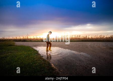 a boy playing in a puddle during sunset on a gravel road Stock Photo