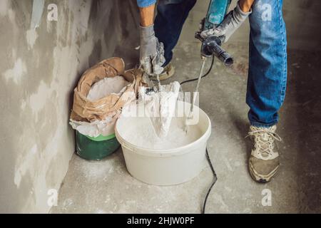 Young male painter kneads putty with water in a bucket using a hand-held mixer for building mixes Stock Photo