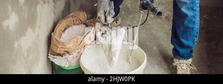 Young male painter kneads putty with water in a bucket using a hand-held mixer for building mixes BANNER, long format Stock Photo