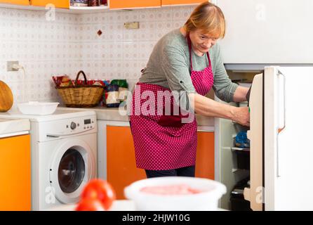 Mature housewife takes food out of the fridge in the kitchen Stock Photo