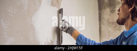 master is applying white putty on a wall and smearing by putty knife in a room of renovating house in daytime BANNER, long format Stock Photo