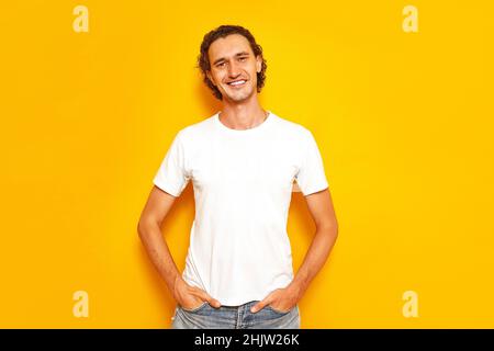 man in casual white T-shirt standing on an isolated yellow background keeps his hands in his pockets Stock Photo