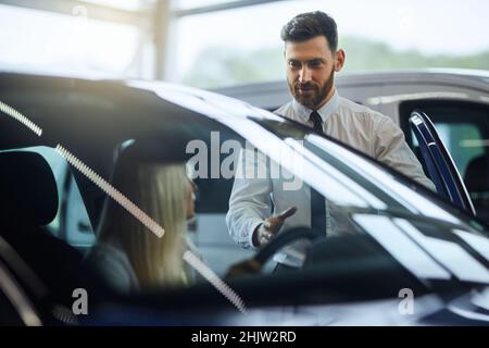 Caucasian salesman showing to female customer new luxury car at showroom. Happy young woman choosing new vehicle at auto salon. Purchase and people concept. Stock Photo