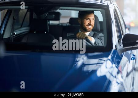 Smiling handsome man visiting modern car salon for choosing new auto. Happy businessman sitting inside luxury transport and testing interior. Stock Photo