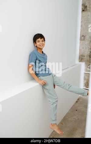 Chill indian boy sitting on a white wall shelf, pressing with his foot against something. Smiling Stock Photo