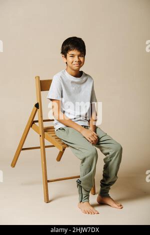 Amused indian boy sitting on a wooden folding chair in a beige room. He is leaning forward with the chair, looking at the camera. Stock Photo