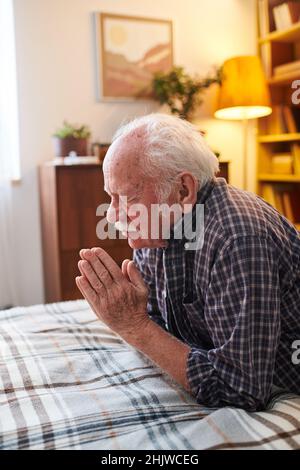 Senior man with his eyes closed sitting in front of the bed and praying before sleep in his room Stock Photo