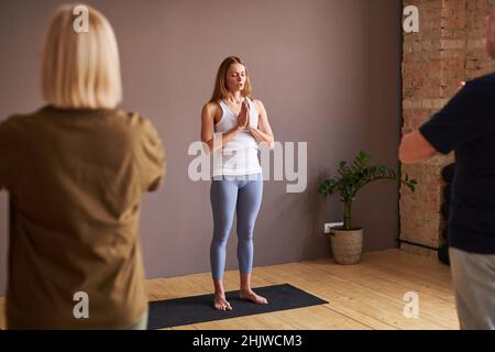 Young fitness instructor practicing yoga with other people during yoga class in gym Stock Photo