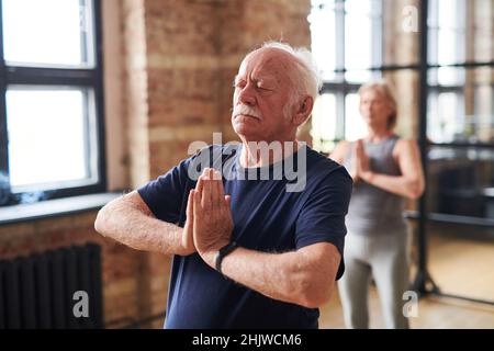Senior man standing in yoga pose with his eyes closed and meditating during class with other people in health club Stock Photo