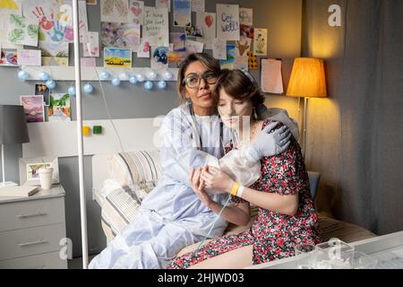 Nurse in protective wear supporting the sick girl during her disease, they sitting on the hospital bed and embracing Stock Photo