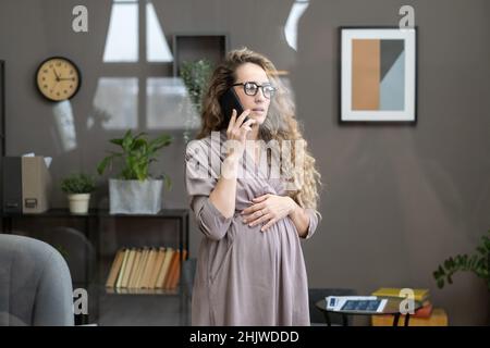 Young blond pregnant businesswoman in smart casualwear speaking on mobile phone while standing in office Stock Photo