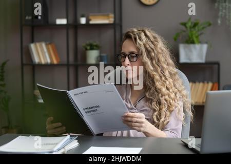 Young serious businesswoman looking through financial documents while sitting by desk against office environment Stock Photo