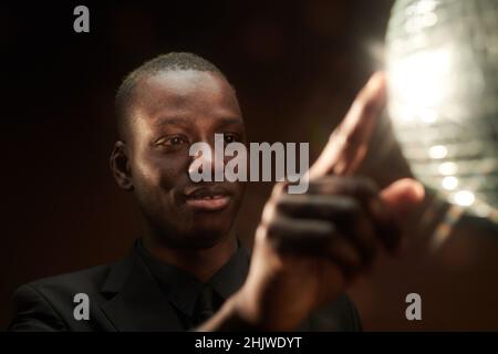 Happy young African man touching discoball and looking at it in the night club while standing in front of camera Stock Photo