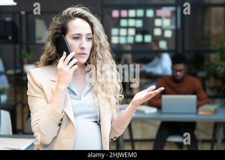 Young confident pregnant businesswoman consulting colleague by mobile phone while standing against coworkers in office Stock Photo