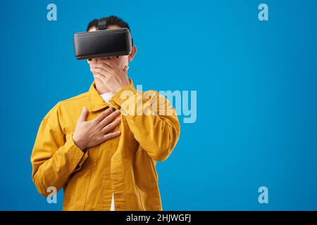 Shocked on scared young man in virtual reality headset covering mouth with hand when looking at something disgusting in videogame Stock Photo
