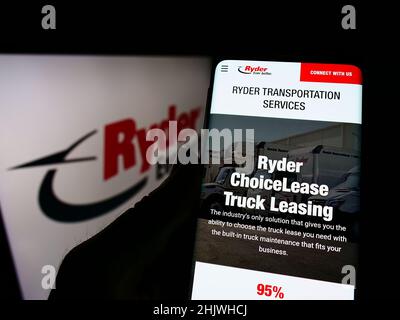 Person holding cellphone with website of US transportation company Ryder System Inc. on screen in front of logo. Focus on center of phone display. Stock Photo