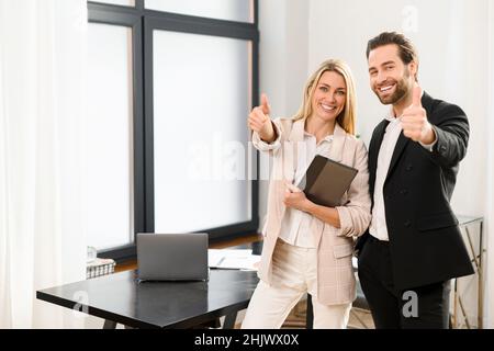 Cool. Young beautiful team of man and woman is using digital tablet while standing at the office. They holding tablet in hands, looking at camera and showing cool gesture Stock Photo