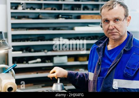 Portrait of adult turner 50-55 years old in glasses and work clothes. Worker in lathe shop looks into camera against background of rack of tools. Copy space. Real scene. Stock Photo