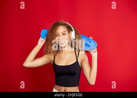 Cheerful African American curly haired female hipster with penny board listening to music in headphones and looking at camera against red background in studio Stock Photo
