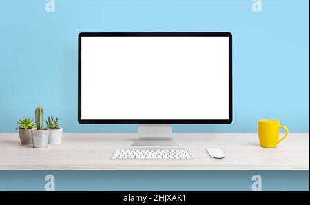 Computer display with white isolated screen for presentation on work desk. Clean composition with coffee mug and plants. Blue wall in background Stock Photo