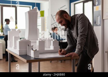 Architect drawing blueprints at desk in modern architectural office next to white foam scale model of skyscraper. Engineer making notes on construction plan leaning over desk. Stock Photo