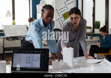 Team of two professional architects working on blueprints on desk in architectural office in front of laptop with construction plans. Colleagues discussing design of white foam building model. Stock Photo