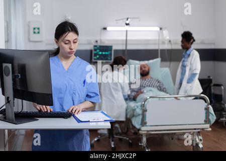 Nurse using personal computer to complete patient admission chart while looking at clipboard with medical history in hospital ward. Team of doctors consulting man while nurse is reading test results. Stock Photo