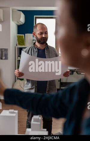 Standing bearded architect looking focused at construction plans collaborating with colleague engineer in modern office. Architects doing architectural design creative teamwork on foam scale model. Stock Photo